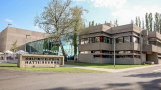 Ein modernes Gebäude mit Glaswänden und roten Rahmen trägt den Namen Kulturzentrum Mattersburg. Vor dem Gebäude steht ein Baum und ein Weg führt zum Eingang.