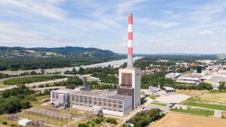 Luftaufnahme eines großen Kraftwerks mit einem hohen Schornstein, umgeben von einer Landschaft mit Bergen, einem Fluss und Bäumen.