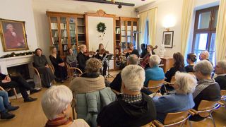 Eine Gruppe von Musikern spielt Violinen und Celli in einem Raum mit Holzstühlen und Regalen. Eine Büste und ein Bilderrahmen sind an der Wand.