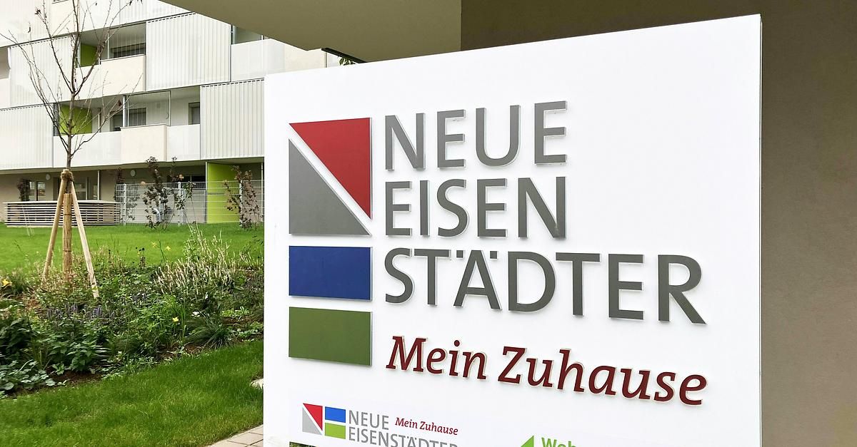Ein weißes Schild mit dem Text 'Neue Eisen Stadter Mein Zuhause' steht vor einem weißen Gebäude mit Balkon und einem grünen Rasen.