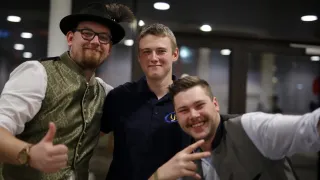 Drei Männer in einem schwach beleuchteten Raum, zwei in traditioneller deutscher Kleidung, einer in einem marineblauen Poloshirt, alle lächelnd und posieren für ein Foto.