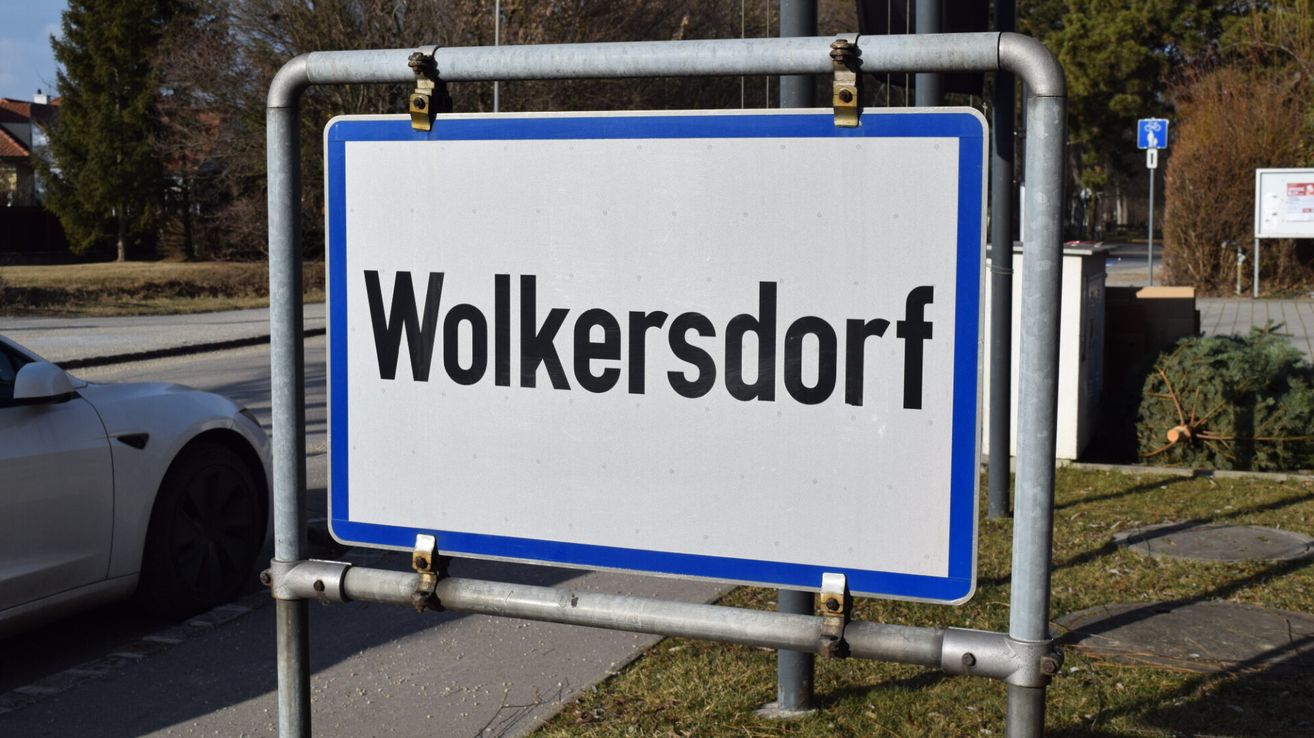 Ein weißes Schild mit schwarzen Buchstaben, die 'Wolkersdorf' lesen, montiert an einem Metallpfosten, mit einem Grasbereich darunter sichtbar.