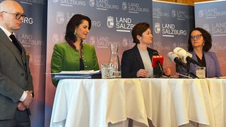 Zwei Frauen in Geschäftskleidung sitzen mit Mikrofonen an einem Tisch vor einem Hintergrund mit Salzburgs Logo und Text.