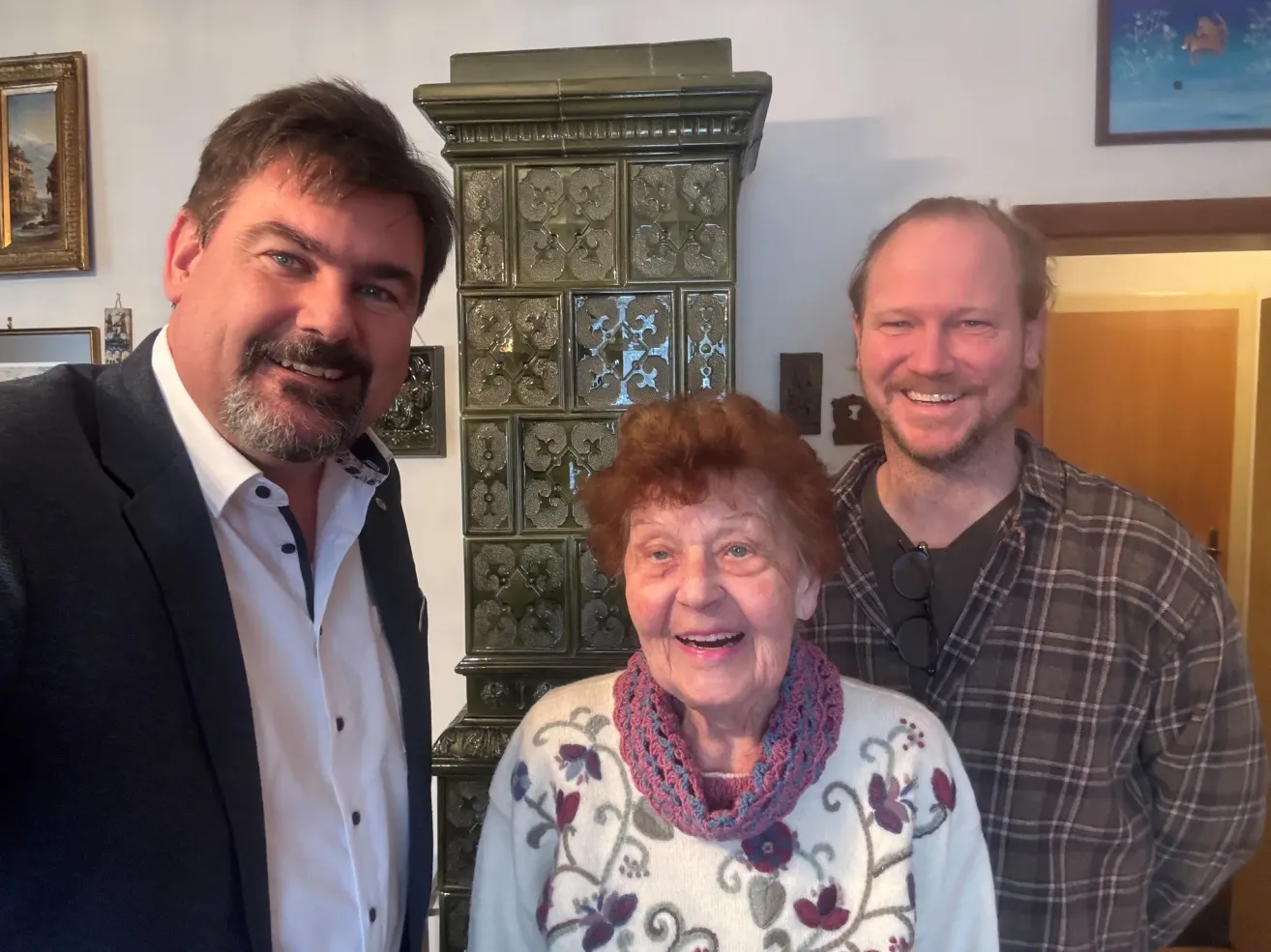 Drei Menschen stehen gemeinsam in einem Raum und lächeln für ein Foto. Eine Frau mit roten Haaren, ein Mann mit Bart und ein weiterer Mann mit Brille. Ein dekorativer Schrank steht hinter ihnen.