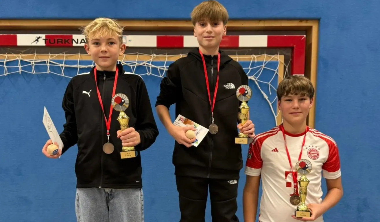 Three boys stand next to each other holding trophies. The boy on the left wears a black jacket and gray pants. The boy in the middle wears a black jacket with a logo and holds a medal. The boy on the right wears a white shirt with red trim. Behind them is a blue wall with a red and white striped design.
