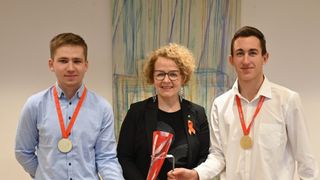 Drei Personen stehen zusammen und lächeln. Die Frau in der Mitte hält eine rot-weiße Trophäe. Beide Männer tragen Medaillen. Der Hintergrund zeigt ein farbenfrohes abstraktes Gemälde.