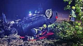 Zwei Feuerwehrleute kümmern sich um eine Person auf einer roten Decke. Ein blaues Auto steht auf dem Kopf auf schlammigem Boden mit einem Rad ab.