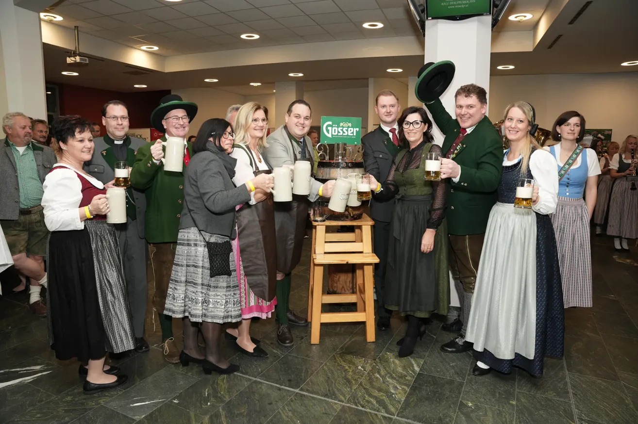 Eine Gruppe von Menschen in traditioneller bayerischer Kleidung hebt Biergläser in einem Saal. Sie stehen um einen Tisch mit einem großen Fass, das mit 'Gösser' beschriftet ist.