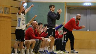 Eine Gruppe von Menschen klatscht in einer Sporthalle. Ein Mann in einer schwarzen Jacke steht mit zusammengeklappten Händen. Es gibt Menschen, die um ihn herum sitzen und stehen. Die Wand ist grün und rechts ist eine Tür.