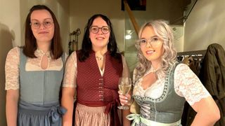 Drei Frauen in traditioneller Kleidung stehen lächelnd nebeneinander. Die mittlere Frau hält ein Weinglas. Sie befinden sich in einem Raum mit einer Treppe im Hintergrund.
