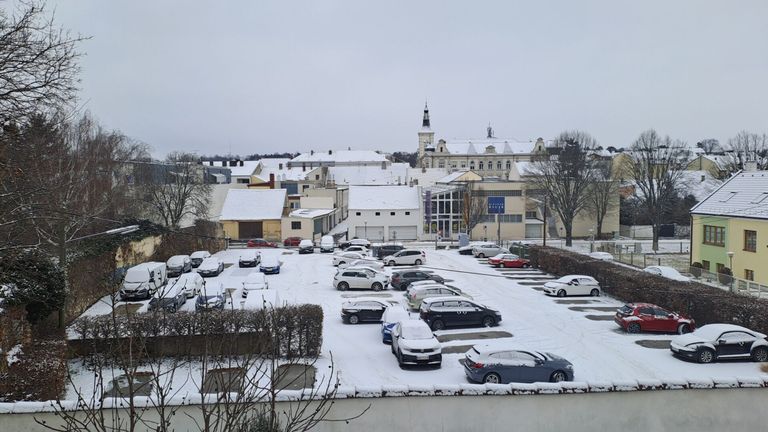 Ein verschneiter Parkplatz mit mehreren geparkten Autos. Schnee bedeckt den Boden, die Bäume und die Gebäude. Ein Gebäude mit einem blauen Schild ist im Hintergrund zu sehen.