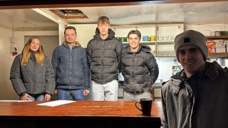 Vier Männer stehen in einem Raum, drei von ihnen tragen Jacken, einer davon bläst Rauch. Dahinter steht ein Regal mit Tassen. Ein Tisch hat ein Papier und eine Tasse.