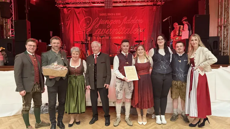 Eine Gruppe von Menschen in traditioneller Kleidung posiert für ein Foto vor einem roten Hintergrund. Ein Mann hält ein Zertifikat, und eine Frau lächelt. Dahinter befindet sich eine Bühne mit Musikinstrumenten.