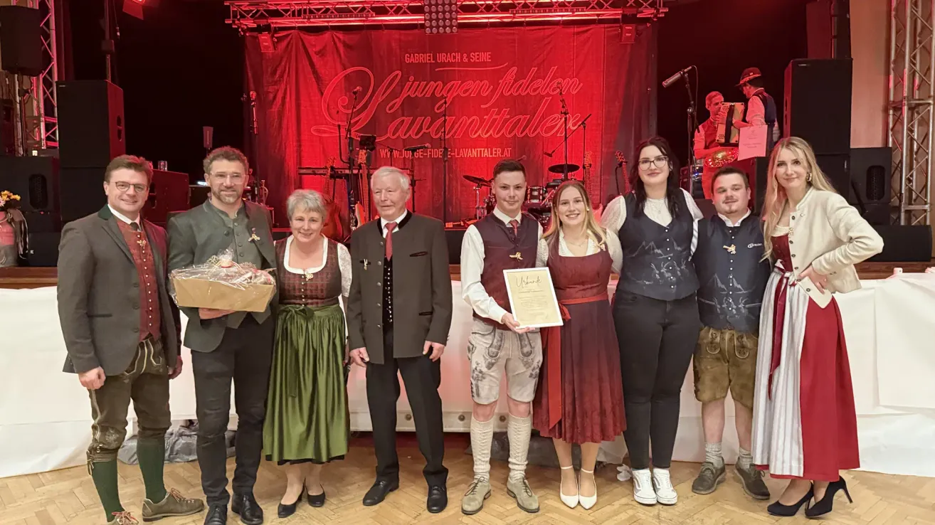 Eine Gruppe von Menschen in traditioneller Kleidung posiert für ein Foto vor einem roten Hintergrund. Ein Mann hält ein Zertifikat, und eine Frau lächelt. Dahinter befindet sich eine Bühne mit Musikinstrumenten.