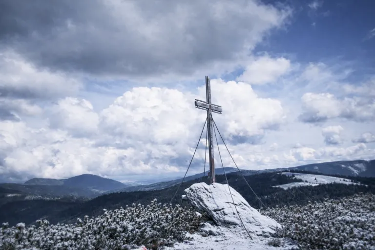 Ein hölzernes Kreuz steht auf einem verschneiten Gipfel mit rauen Bergen und einem bewölkten Himmel im Hintergrund.