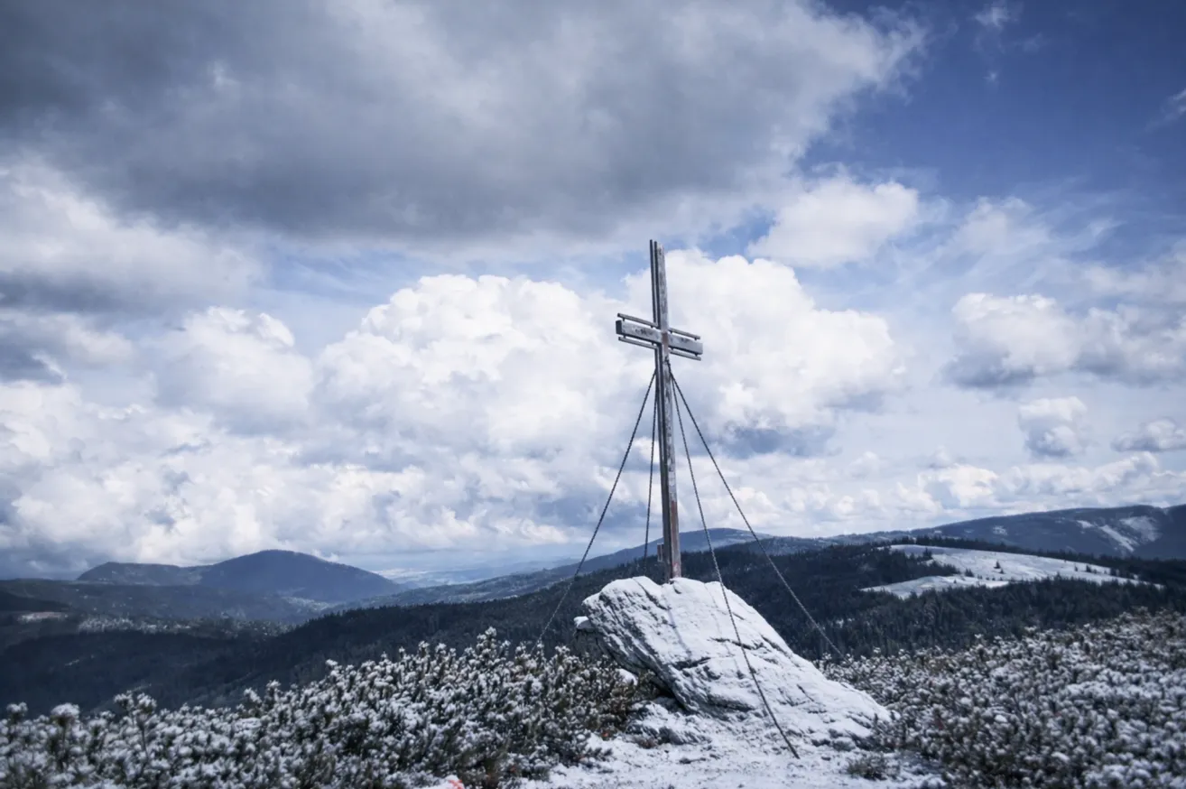 Ein hölzernes Kreuz steht auf einem verschneiten Gipfel mit rauen Bergen und einem bewölkten Himmel im Hintergrund.