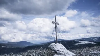 Ein hölzernes Kreuz steht auf einem verschneiten Gipfel mit rauen Bergen und einem bewölkten Himmel im Hintergrund.