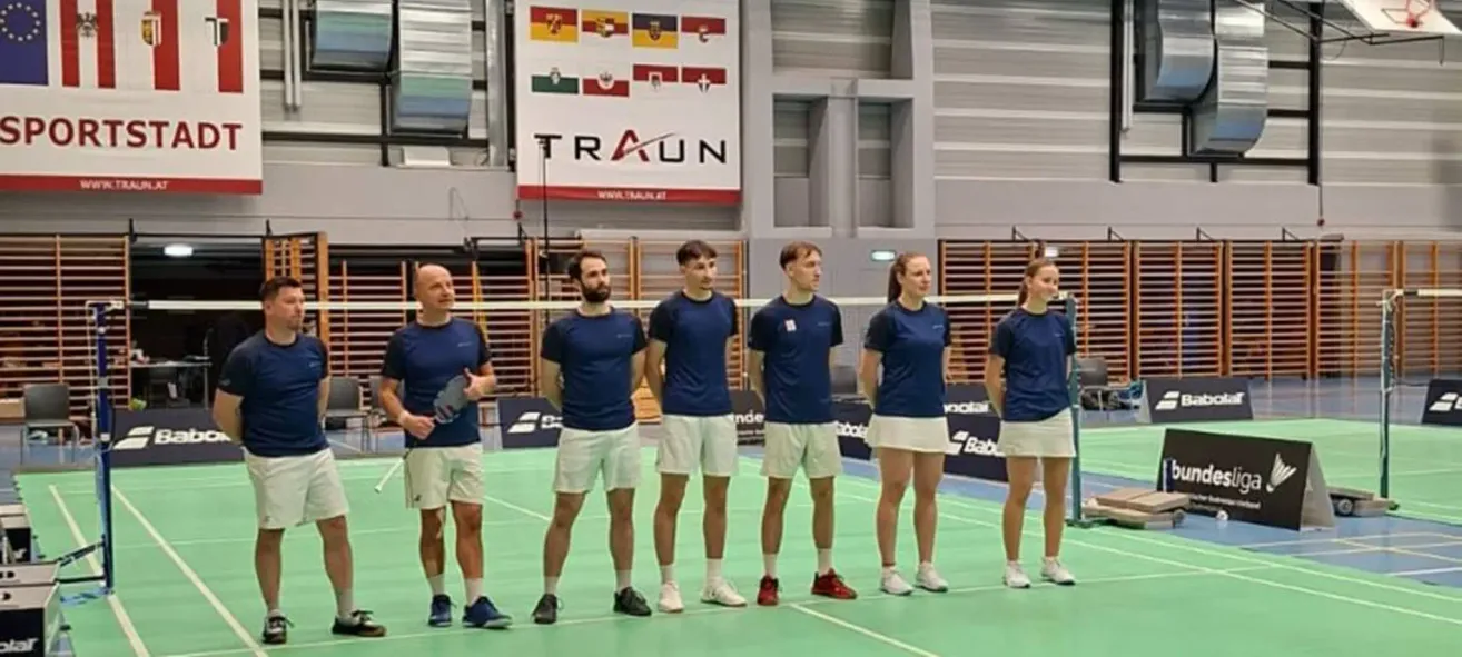 Ein Badmintonteam in blauen Uniformen steht auf einem Spielfeld. Dahinter ist ein Banner mit mehreren Flaggen und dem Wort Traun zu sehen.
