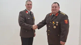 Zwei Männer in Militäruniformen schütteln sich die Hände in einem Raum mit weißer Wand und Holzboden.