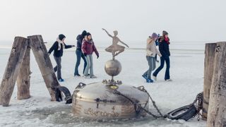 Eine Gruppe von Menschen geht auf dem Eis in der Nähe einer Metallstruktur. Die Struktur hat eine Statue einer Ballerina oben drauf. Sie tragen Winterkleidung. Das Wetter ist neblig.
