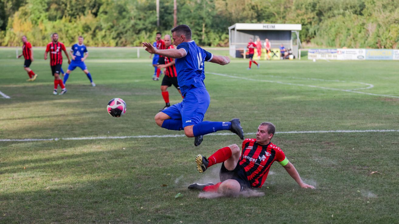 Fußballspieler befinden sich auf einem Feld. Ein Spieler in Blau springt, um den Ball zu schießen, während ein anderer in Rot auf dem Boden liegt.