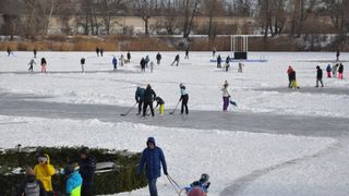 Menschen eislaufen und spielen Eishockey auf einem gefrorenen See, umgeben von Bäumen und einem entfernten Gebäude. Einige gehen, andere bereiten sich auf Aktivitäten vor.