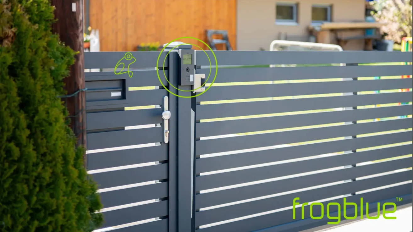 Ein graues Metalltor mit einem grünen Kreis um eine digitale Verriegelung und Tastatur. Hinter dem Tor steht ein Holzhaus mit einem Fenster.