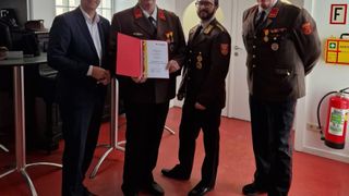 Vier Männer in formeller Kleidung stehen in einem Raum. Der Mann in der Mitte hält eine rote Mappe. Zwei andere tragen Militäruniformen. Der Mann auf der linken Seite trägt einen Anzug.
