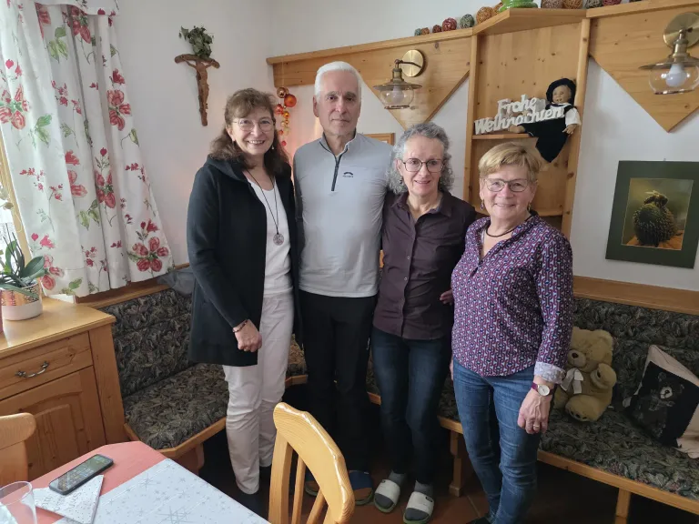 Vier Menschen lächeln und posieren für ein Foto im Wohnzimmer. Die Frau links trägt eine schwarze Jacke und weiße Hosen. Der Mann in der Mitte trägt ein weißes Hemd. Die Frau neben ihm trägt ein lila Hemd. Die Frau rechts trägt ein blaues Blumenhemd und eine Uhr.