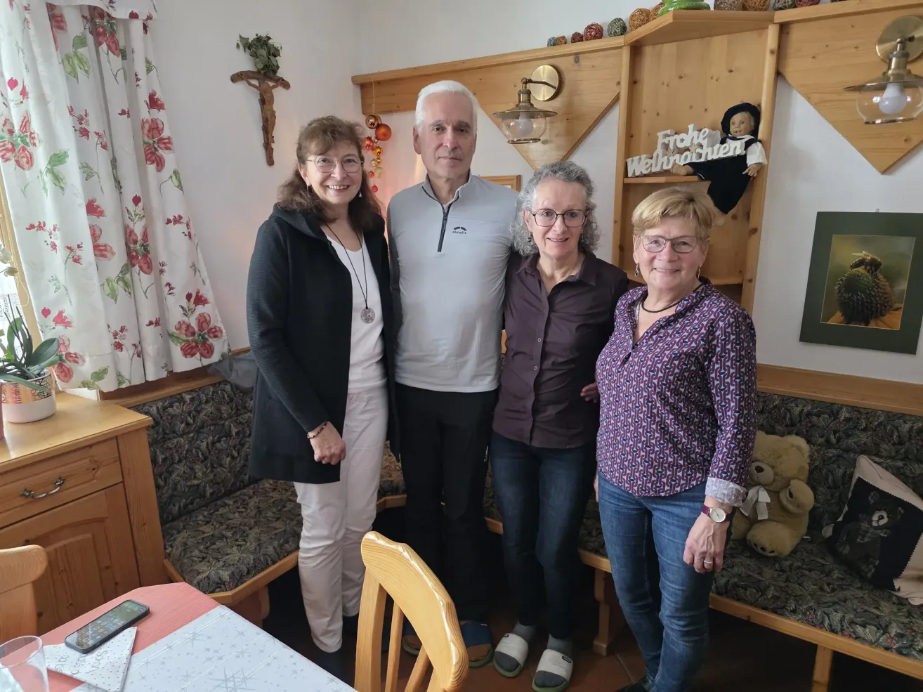 Vier Menschen lächeln und posieren für ein Foto im Wohnzimmer. Die Frau links trägt eine schwarze Jacke und weiße Hosen. Der Mann in der Mitte trägt ein weißes Hemd. Die Frau neben ihm trägt ein lila Hemd. Die Frau rechts trägt ein blaues Blumenhemd und eine Uhr.