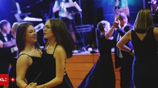 Zwei Frauen in schwarzen Kleidern tanzen auf einer Bühne, während eine Band im Hintergrund spielt. Eine Frau trägt eine Brille und eine Halskette.