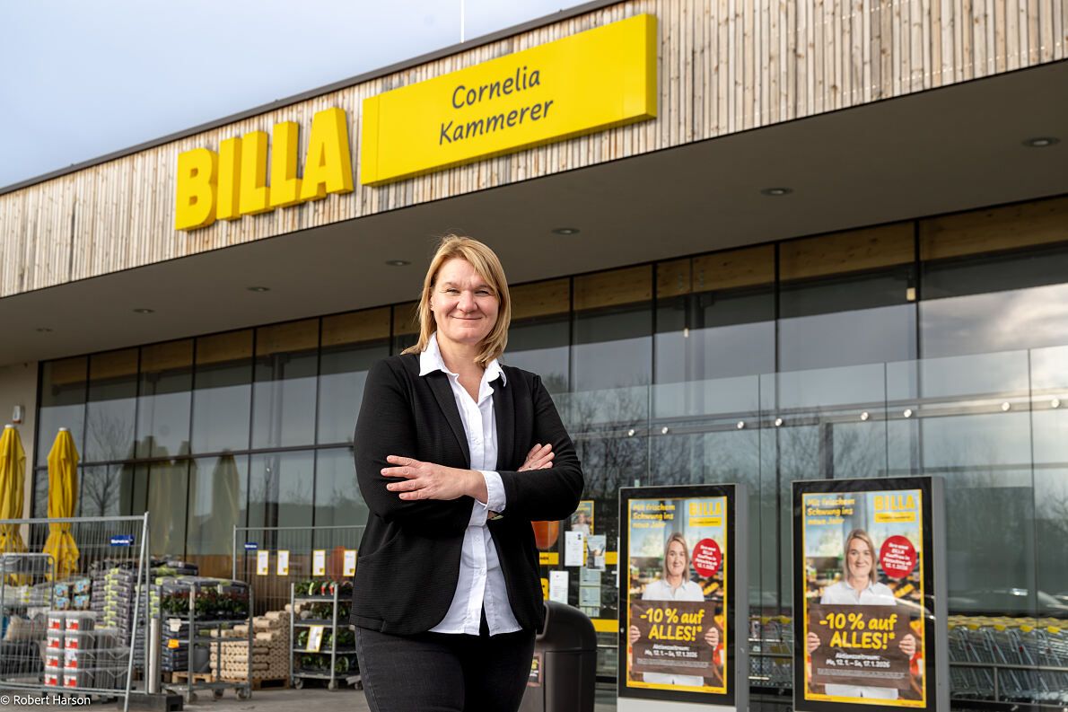 Eine Frau steht selbstbewusst vor einem BILLA-Laden, lächelt und hat die Arme verschränkt. Der Laden hat eine moderne Holz- und Glasfassade mit einem großen gelben Schild, das 'CORNELIA KAMMERER' besagt. Hinter ihr befinden sich Außendisplays und ein Mülleimer.