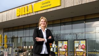 Eine Frau steht selbstbewusst vor einem BILLA-Laden, lächelt und hat die Arme verschränkt. Der Laden hat eine moderne Holz- und Glasfassade mit einem großen gelben Schild, das 'CORNELIA KAMMERER' besagt. Hinter ihr befinden sich Außendisplays und ein Mülleimer.