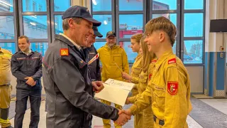 Ein Mann in Uniform überreicht einem lächelnden Jugendlichen in gelber Feuerwehruniform ein Zertifikat. Hinter ihnen beobachten andere Feuerwehrleute, und eine Glaswand zeigt einen Feuerwehrwagen.