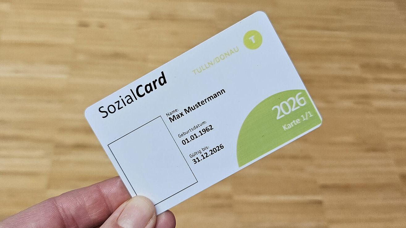 Eine weiße SozialCard wird in der Hand gehalten, mit dem Namen Max Mustermann, geboren am 01.01.1962. Die Karte ist gültig bis 31.12.2026. Sie hat einen grünen Kreis mit der Nummer 2026 und dem Text Karte 1/1.
