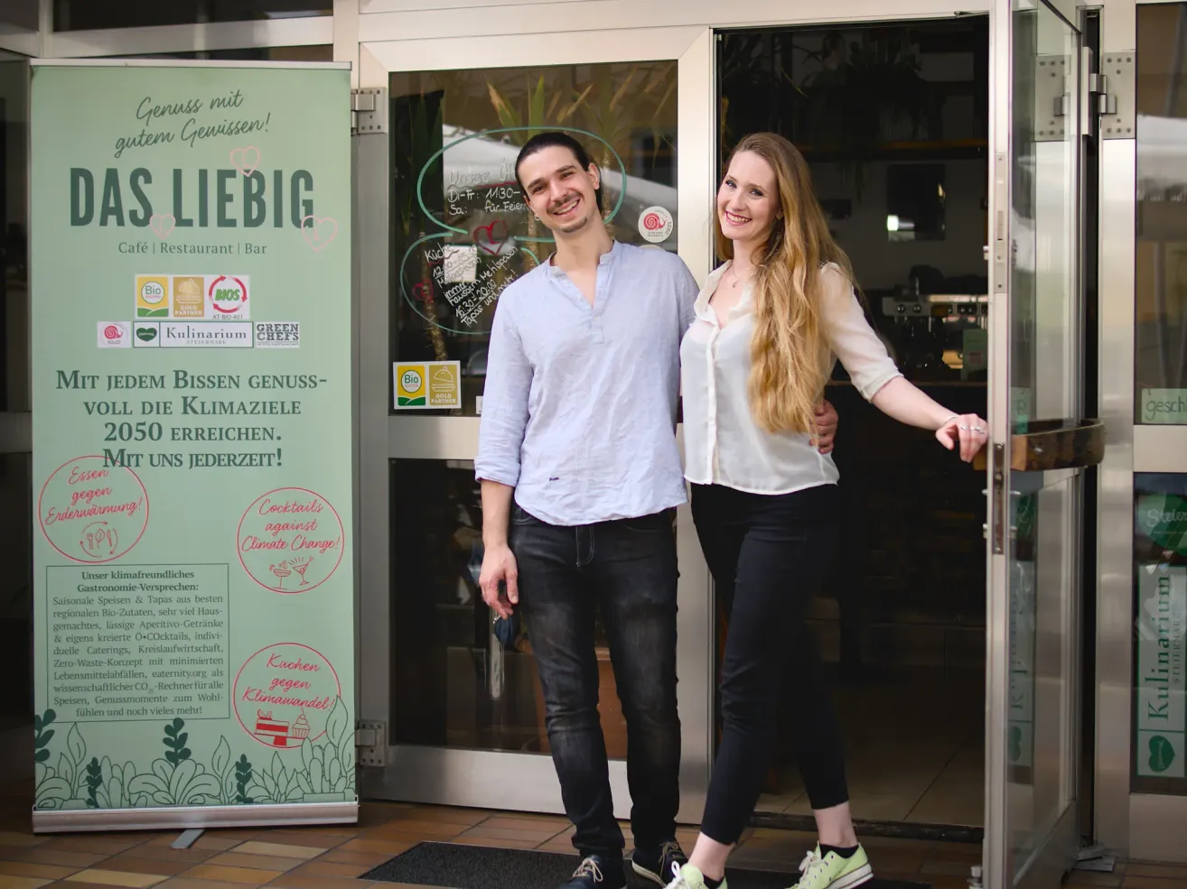 Ein Mann und eine Frau stehen lächelnd vor einem Restaurant und machen ein Foto. Das Restaurant hat ein Schild mit den Worten 'Liebig' und 'Restaurant Bar'. Der Mann trägt ein hellblaues Hemd und schwarze Hose, während die Frau ein weißes Blusenkleid und schwarze Hose trägt.