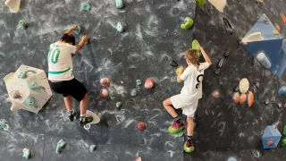 Zwei Kinder klettern an einer Kletterwand mit farbigen Griffen. Eines trägt ein Shirt mit der Nummer 10, das andere hat die Nummer 5. Sie benutzen Kletterschuhe mit grünen Griffen.