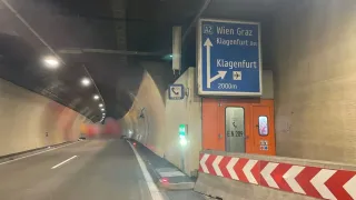 Der Tunneleingang zeigt ein blaues Schild mit einem Pfeil, der auf Klagenfurt zeigt. Das Schild zeigt Klagenfurt Ost und 2000m. Eine orange Tür mit der Nummer 209 ist sichtbar.