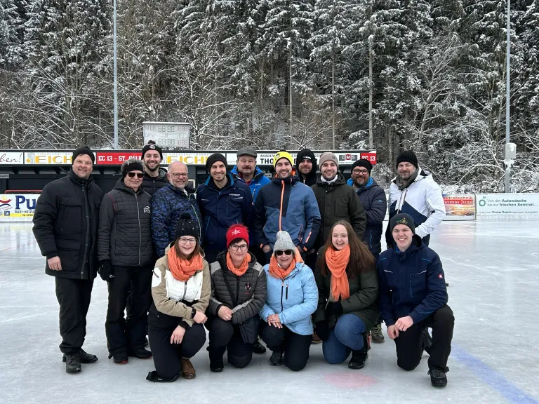 Eine Gruppe von Menschen posiert für ein Foto auf einer Eisbahn. Sie sind alle in Winterkleidung gekleidet. Hinter ihnen befindet sich ein Zaun mit Werbetafeln. Die Bäume im Hintergrund sind mit Schnee bedeckt.