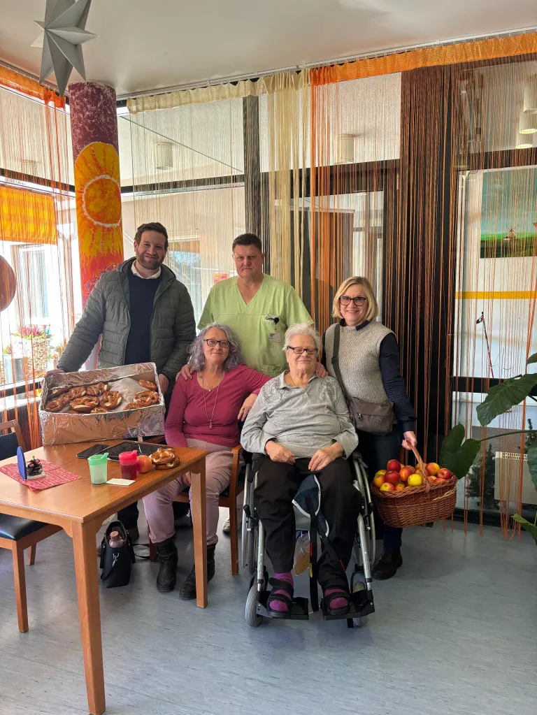 Fünf Menschen posieren für ein Foto in einem Raum. Zwei Frauen sitzen auf einem Stuhl, eine von ihnen ist im Rollstuhl. Sie alle lächeln. Ein Tisch mit Essen und einem Korb mit Äpfeln steht vor ihnen.