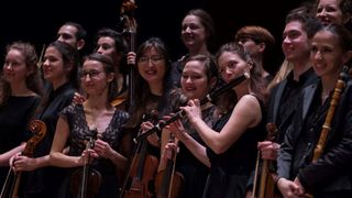 Eine Gruppe von Musikern, darunter Geiger, Cellisten und eine Flötistin, lächelt und posiert für ein Foto auf der Bühne.