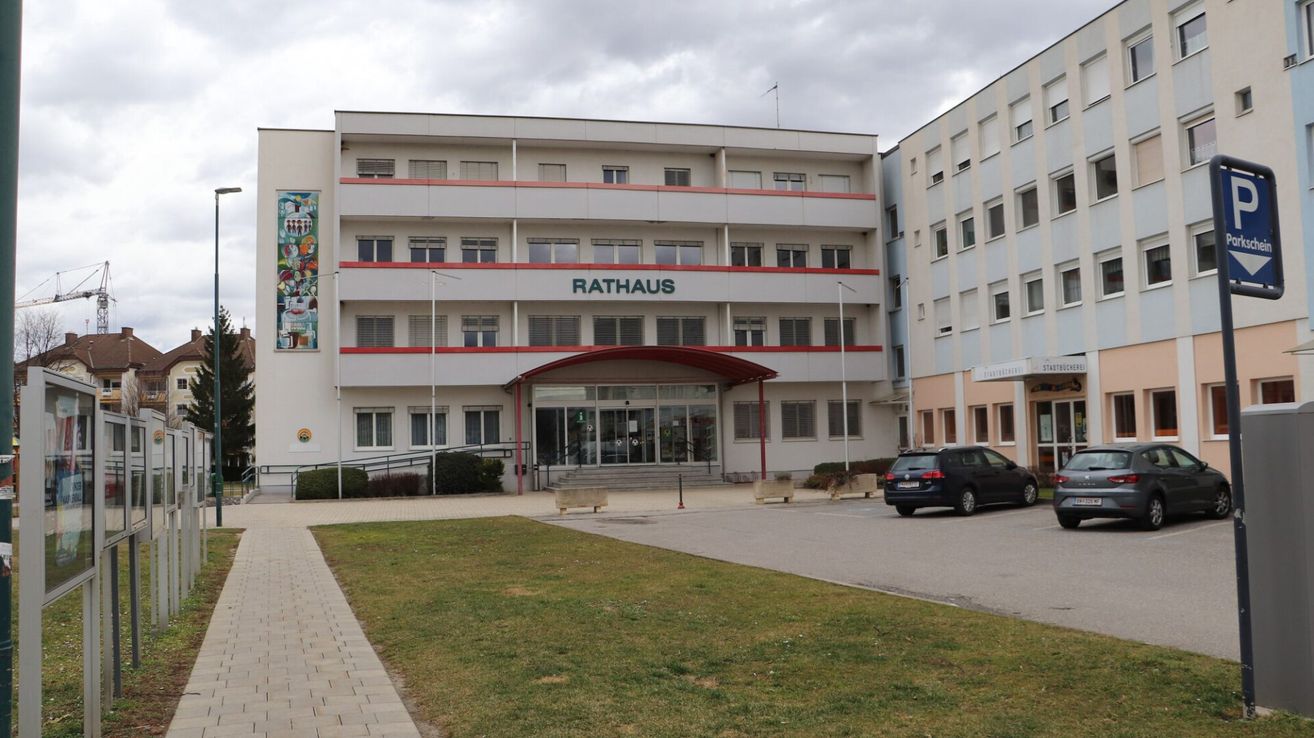 Ein weißes Gebäude mit dem Namen Rathaus über dem Eingang, einem roten Dach über dem Eingang und einem großen Fenster links. Ein schwarzes Auto ist vor dem Gebäude geparkt.
