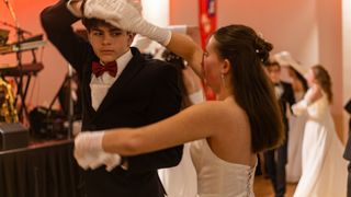 Ein junger Mann in einem schwarzen Anzug und einer roten Schleife wird von einer Frau in einem weißen Kleid unterstützt. Sie tragen beide weiße Handschuhe.