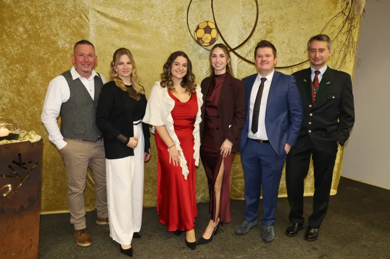 Eine Gruppe von sechs Personen, darunter vier Frauen und zwei Männer, posiert für ein Foto vor einer goldenen Wand mit einem Fußballdesign.