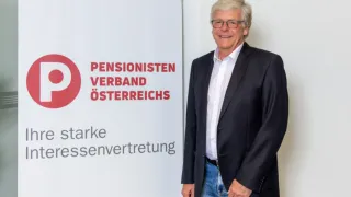Ein älterer Mann steht lächelnd vor einem Pensionisten Verband Osterreichs Banner. Er ist in einem schwarzen Anzug und Jeans gekleidet.