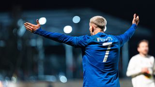 Ein männlicher Fußballspieler feiert ein Tor mit erhobenen Armen. Er trägt ein blaues Trikot mit der Nummer 7. Der Hintergrund ist dunkel mit hellen Scheinwerfern.