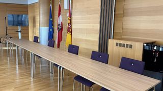 Ein leerer Konferenzraum mit einem langen Holztisch, lila Stühlen und drei Flaggen an der Wand. Die Wand besteht aus Holz.
