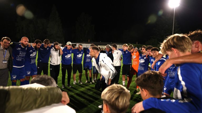 A soccer team in blue and white uniforms, including a man in white bending over, is gathered together at night.