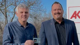 Zwei Männer stehen vor einem weißen Schild mit einem roten Buchstaben A und lächeln. Der Mann auf der linken Seite hält eine kleine Karte. Der Mann auf der rechten Seite trägt einen dunklen Anzug und ein schwarzes Hemd. Sie sind draußen.