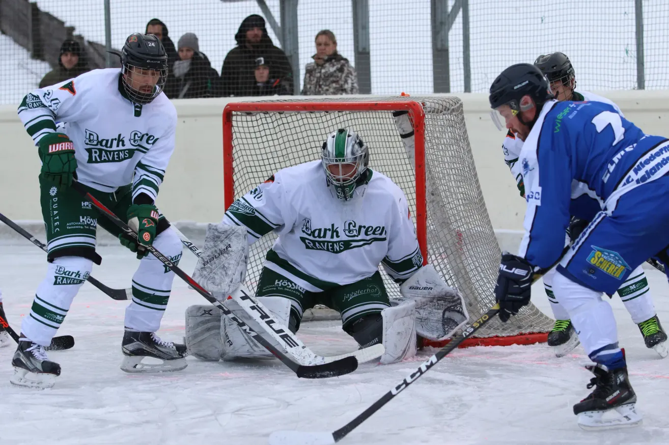 Eishockeyspieler in Uniform fahren auf dem Eis. Ein Spieler hockt vor dem Tornetz, während ein anderer Spieler auf das Tor zielt. Zuschauer in Winterkleidung beobachten von einer Umzäunung aus.